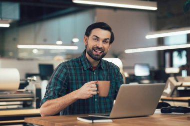 İş yerindeki masada elinde kupayla oturmuş laptopun önünde gülümseyen kareli gömlekli olumlu bir adam.