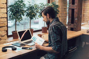 Laptop ekranına bakıp bir kağıt üzerinde grafik tutan konsantre genç bir çalışan. Yanında akıllı telefon kulaklıkları ve defter vardı.