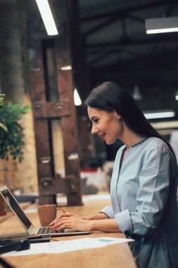Koyu renk saçlı, gülümseyen ve dizüstü bilgisayarda yazarken bilgisayarın ekranına bakan güzel bayan.