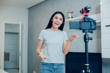 Tutkulu, kendinden emin, günlük kıyafetler giymiş genç bir kadın kameranın önünde duruyor ve konuşurken el kol hareketi yapıyor.