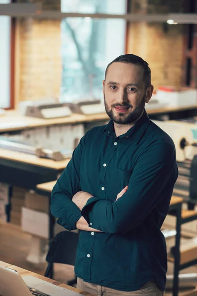 Handsome employee of a publishing house standing with crossed arms in front of the table with laptop on it