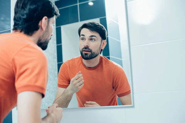 鏡の前のバスルームに立つオレンジのTシャツの魅力的な若い男と彼の反射を見て — ストック写真