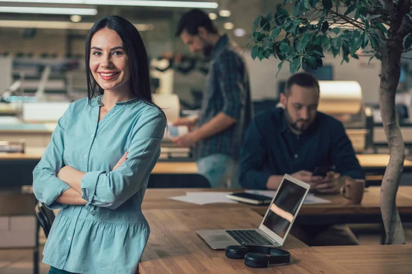 Cheerful beautiful lady standing with crossed arms by the table while being in the office with laptop behind her back. Website banner
