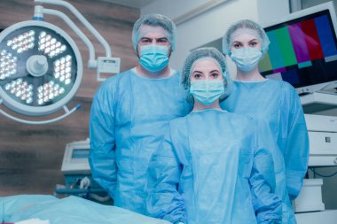 Bir erkek ve iki kadın doktor temiz mavi üniformalı ameliyathanede bir lambanın altında bekliyorlar.