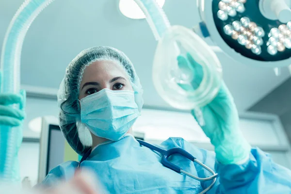Tıbbi maskeli ve koruyucu elbiseli bir doktor elinde anestezi maskesiyle ayakta duruyor.