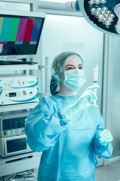 Professional medical worker standing under the lamp in modern operating room and looking at the medication in a syringe