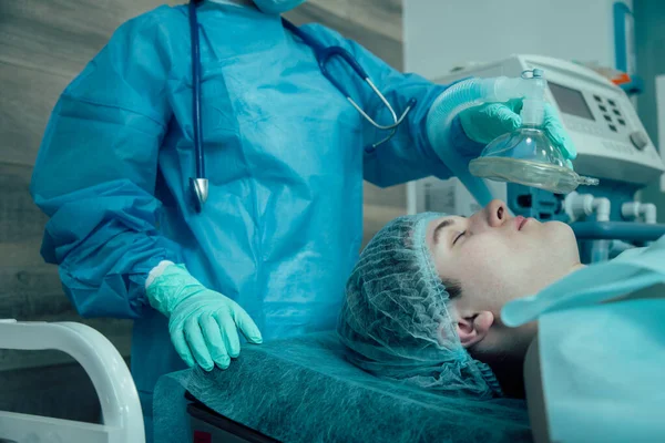 Ameliyathanede bir masada uzanan genç bir yetişkin ve yüzünde anestezi maskesiyle profesyonel bir doktor.