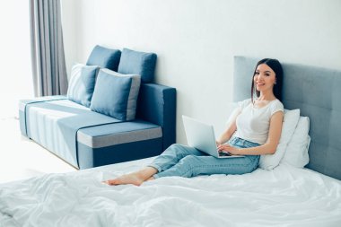 Kucağında dizüstü bilgisayarla yatakta rahat rahat oturan ve gülümseyen neşeli, yalınayak bir kadın.