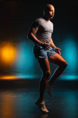 Koşu yaparken ciddi görünen şortlu ve tişörtlü cesur bir genç. Arkaplanda turuncu deniz mavisi ışığı