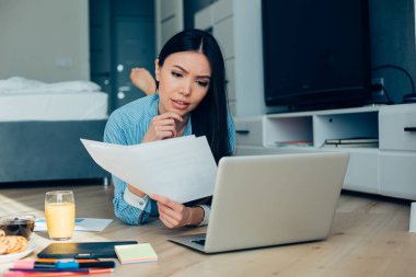Güzel bir kadın yere uzanmış, yanında kırtasiye malzemeleri ve meyve suyuyla dizüstü bilgisayar ekranına bakarken kaşlarını çatıyor.