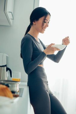 Mutfakta elinde bir kase ve mutfak dolabının yanında kaşıkla duran güzel bir kadın.