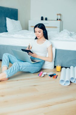 Genç bir kadın yerde yatağın yanında oturuyor ve çizim tahtası kullanırken dizüstü bilgisayara bakıyor.