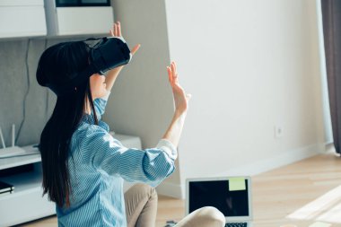 Uzun saçlı bir kadın dizüstü bilgisayarın önünde oturmuş VR gözlük takarken havaya dokunuyor.