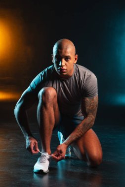 Stüdyoda turuncu deniz mavisi ışığına karşı tek dizinin üstünde oturan ve ayakkabı bağcıklarını bağlayan düşünceli genç adam.