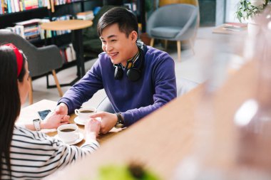 Neşeli genç bir adam ve bir bayan kahve fincanlarıyla masada oturuyor ve el ele tutuşuyorlar.