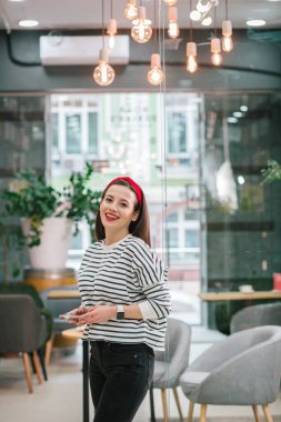 Çizgili bir kazak içinde neşeli, genç bir bayan elinde akıllı bir telefonla cam kapının yanında dururken gülümsüyor.