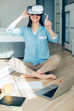 Yerde dizüstü bilgisayarıyla oturan ve sanal gerçeklikteyken görünmeyen bir şeye işaret eden memnun genç bir kadın.