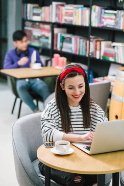 Neşeli, uzun saçlı bir kadın bir fincan kahveyle masada oturuyor ve nazik bir gülümsemeyle ekrana bakıyor.