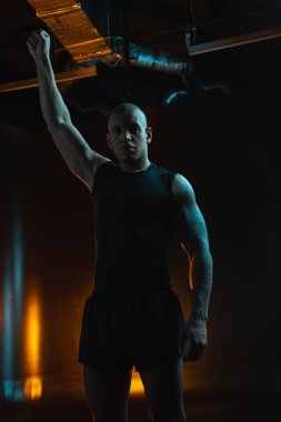 Ciddi sportif kaslı bir adam karanlıkta tek başına duruyor. Yumruğunu kaldırıp mesafeye bakıyor.