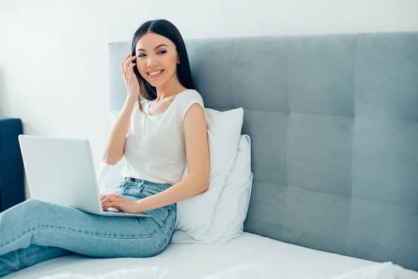 Contented attractive young woman smiling and touching her temple while sitting in a bed with her laptop. Template banner