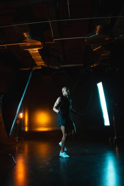Fit young man in the illuminated studio jumping on a skipping rope. Orange light on the background