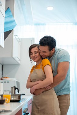 Mutfakta güzel, gülümseyen kız arkadaşının arkasında duran ve onu kucaklayan pozitif genç adam.