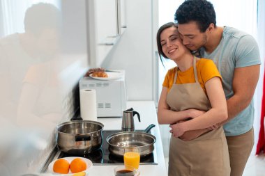 Mutfakta önlük takan genç bir bayan, sevgi dolu erkek arkadaşı onu kucaklarken gözleri kapalı gülümsüyor.