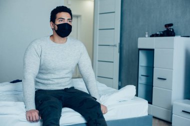 Gündelik giysiler içinde yakışıklı bir adam ve yüzünde siyah bir tıbbi maske var. Yatakta oturuyor ve mesafeye bakıyor.