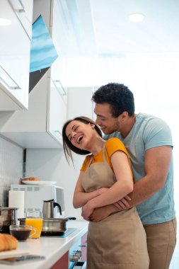 Yakışıklı genç adam mutfakta duruyor ve eğlenen kız arkadaşına sarılırken gülümsüyor.
