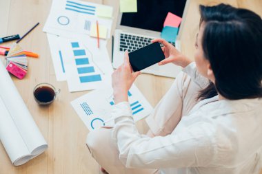 Yerde akıllı telefonuyla oturan genç bir kadın. Önündeki grafiksel çizimler ve renk paletleri