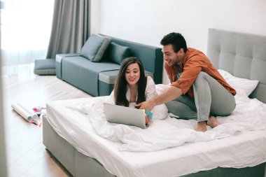 Neşeli genç adam bir yatakta oturuyor ve gülümseyen kız arkadaşı ona bakarken modern bir bilgisayarın ekranını işaret ediyor.