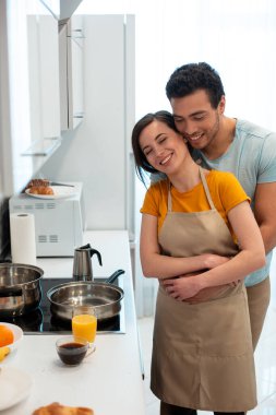 Yakışıklı genç adam, kız arkadaşının arkasında duruyor ve fırının yanında dururken onu kucaklıyor.