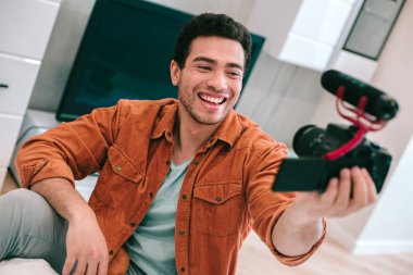 Kendinden emin, rahat giysiler içindeki genç bir adam evde kamerayla oturuyor ve kendini kameraya kaydederken gülümsüyor.