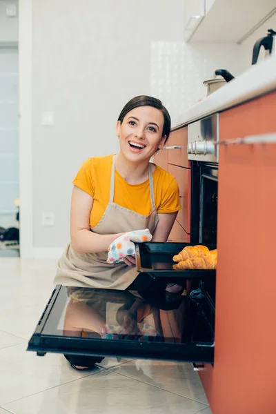 Mirthful Caucasian lady in t-shirt and apron smiling while taking a baking pan with tasty croissants from the oven