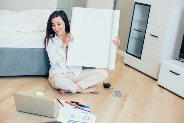 Positive designer showing the roll of engineer paper in her hands while sitting on the floor of her bedroom during the online meeting
