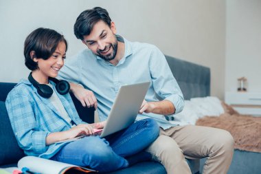Gülümseyen oğluyla kanepede oturan ve dizlerindeki dizüstü bilgisayarı gösteren neşeli dost bir adam.