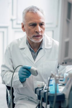 Beyaz önlüklü, elinde ultrason dönüştürücüsüyle oturup ekrana bakan olgun bir doktor.