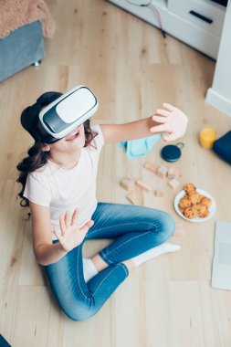 Yüzünde sanal gerçeklik aygıtıyla yerde oturan ve ellerini kaldıran bir çocuğun üst görüntüsü.