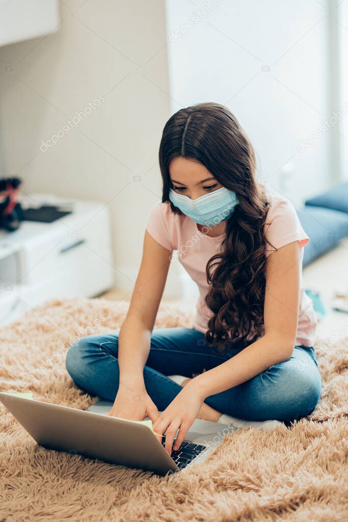 Calm concentrated girl in a medical mask typing on a laptop keyboard while sitting on the bed in her room