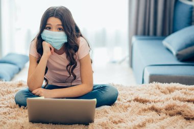 Medikal maskeli şirin bir kız kendini izole bir şekilde dizüstü bilgisayarın önünde otururken çenesini yumruğa koyuyor. Şablon pankartı