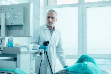 Ciddi bir erkek doktor elinde endoskopla hastasının yanında duruyor ve monitöre bakıyor.