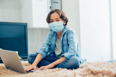 Tıbbi maskeli sakin bir çocuk, önünde modern bir dizüstü bilgisayarla evde dinleniyor.