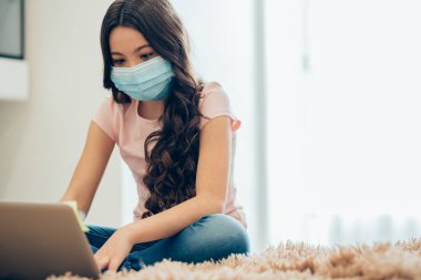 Evde tıbbi maske takan sakin bir kız ve önündeki dizüstü bilgisayara bakıyor. Web sitesi afişi