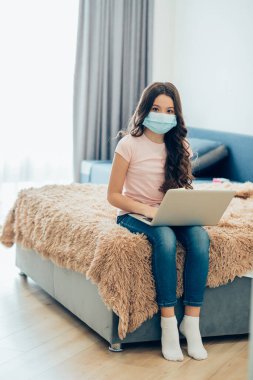 Karantina sırasında dizüstü bilgisayarla yatakta otururken tıbbi maske takan sakin bir çocuk.