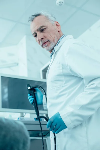 Calm male doctor with an endoscope in hands turning to his patient. Monitor of medical apparatus on the background