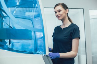 Koyu mavi üniformalı neşeli bayan bir bağışıklık kimyası çözümleyicisinin önünde dururken gülümsüyor.