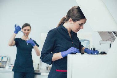 Dikkatli bir laboratuvar uzmanı biyokimyasal analiz cihazına test tüpleri koyuyor ve meslektaşı da arka planda bir pipet makinesi kullanıyor.