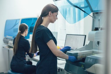 Bağışıklık sistemi analizcisine test tüpü yerleştirirken gülümseyen uzun saçlı kadın laboratuvar uzmanı.