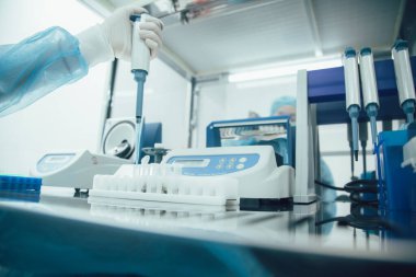 Masanın üzerinde test tüpü rafının kesilmiş fotoğrafı ve plastik eldivenli bir laboratuvar çalışanı elinde otomatik bir pipet makinesi tutuyor.