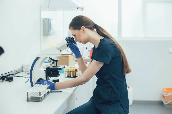 Rolig Langhåret Laboratorietekniker Kigger Mikroskopet Mens Sidder Ved Bordet - Stock-foto